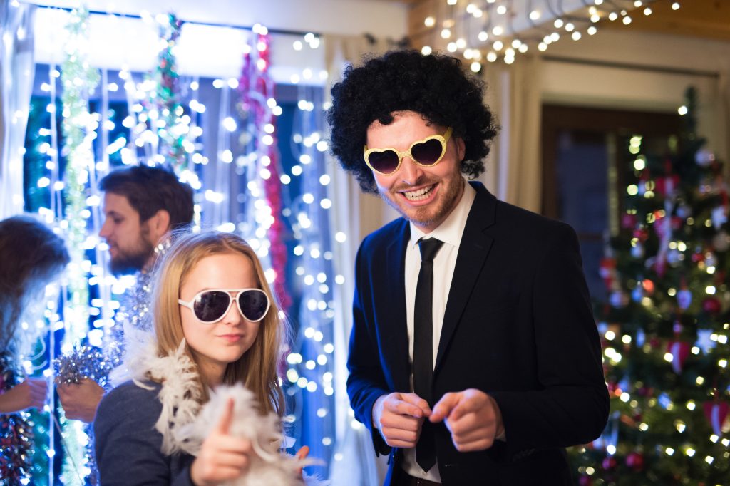Beautiful hipster friends with sunglasses, wig and feather boa celebrating the end of the year, having party on New Years Eve, dancing, chain of lights and Christmas tree behind them.